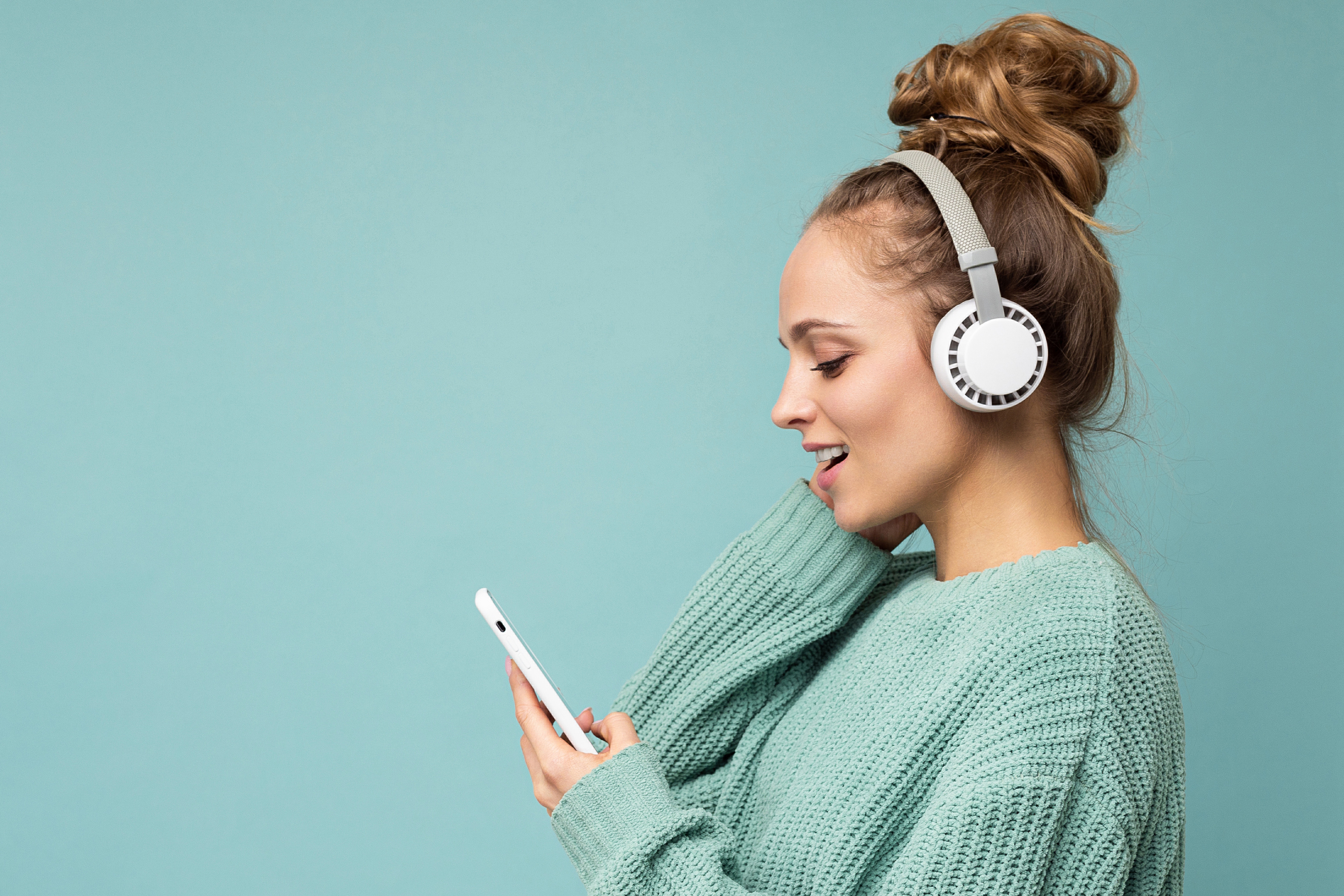 Side-Ptofile Closeup Photo of Beautiful Young Woman Wearing Stylish Casual Outfit Isolated over Colorful Background Wall Wearing White Wireless Headphones and Listening to Music and Using Mobile Phone Messaging Sms Looking at Smartphone Display